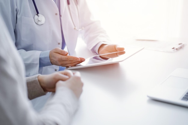 Doctor and patient sitting at the table in clinic. The focus is on female physician's hands using tablet computer, close up. Medicine and healthcare concept.