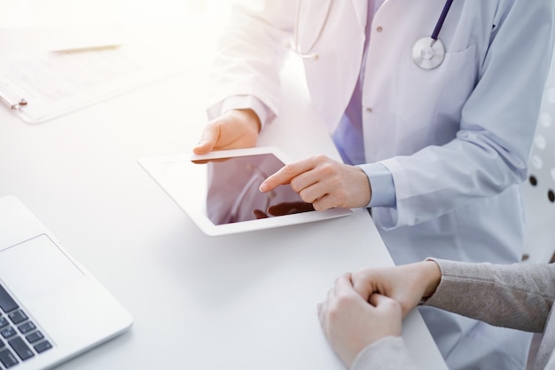 Doctor and patient sitting at the table in clinic. The focus is on female physician's hands using tablet computer, close up. Medicine and healthcare concept.