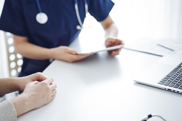 Doctor and patient sitting at the table in clinic while discussing something and using tablet computer. The focus is on female patient's hands, close up. Medicine concept.