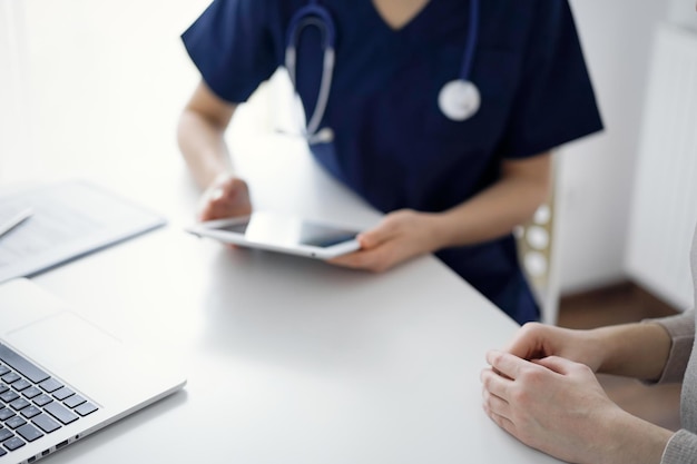 Doctor and patient sitting at the table in clinic while discussing something and using tablet computer. The focus is on female patient's hands, close up. Medicine concept