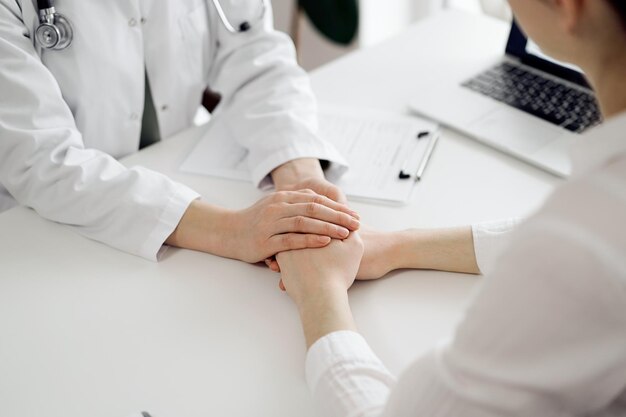 Doctor and patient sitting at the white table near window in clinic. Female physician's hands reassuring woman. Medicine concept