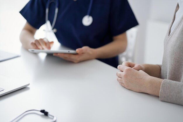 의사와 환자가 앉아서 태블릿 컴퓨터를 사용하는 동안 클리닉에서 서로 이야기하고 닫습니다. 의학 개념