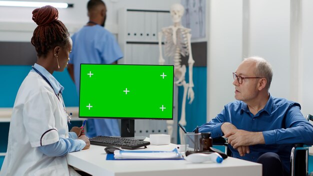 Doctor and patient with impairment using greenscreen on monitor at checkup visit. specialist and old man looking at isolated mockup template with chroma key copyspace. tripod shot