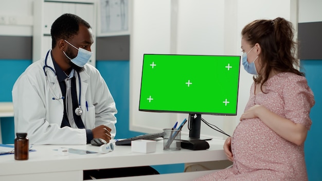 Doctor and patient with pregnancy belly looking at greenscreen on computer during covid 19 pandemic. Isolated chroma key background with mockup template and blank copy space on monitor.