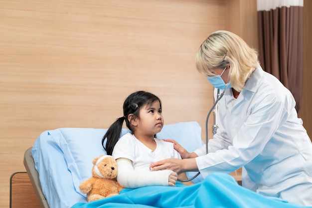 Doctor pediatrician examining a little girl patient on bed with teddy bear by stethoscope