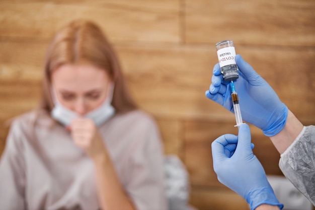 Doctor preparing to vaccine female patient at home. use for prevention, immunization and treatment from corona virus infection. close-up focus on vaccine bottle. female coughing in the background