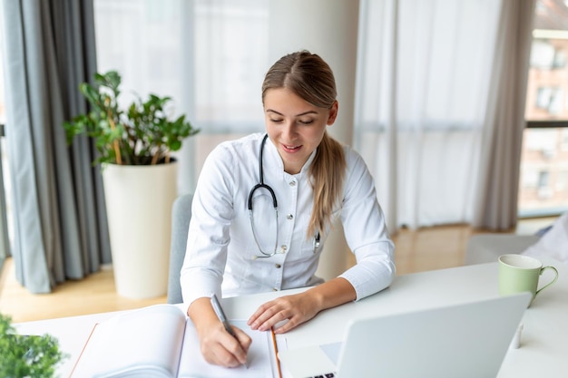 Doctor professional female doctor wearing uniform taking notes\
in medical journal filling documents patient illness history\
looking at laptop screen student watching webinar