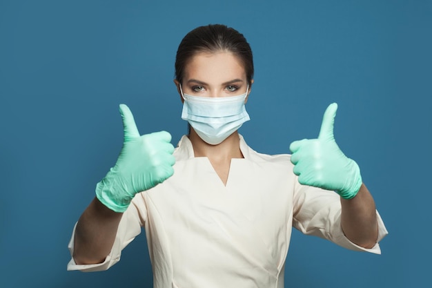 Doctor in professional uniform and protective mask holding thumb up on blue background Medicine safety vaccination and virus covid19 protection concept