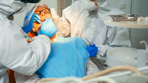 구강 클리닉에서 covid-19 전염병 동안 노인 여성의 치아를 검사하는 보호복을 입은 의사. 안면 보호 마스크와 장갑을 끼고 환자와 이야기하는 램프를 켜는 치열 교정 의사