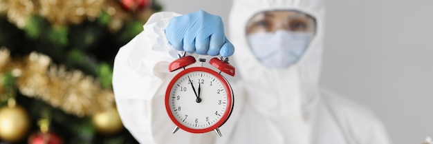 Doctor in protective antiplague suit holding alarm clock near christmas tree remote new years