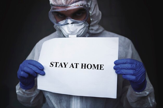 A doctor in a protective biochemistry suit and mask with a paper with inscription stay at home holding on hand on a dark black background. protect from Coronavirus or Covid-19 epidemic