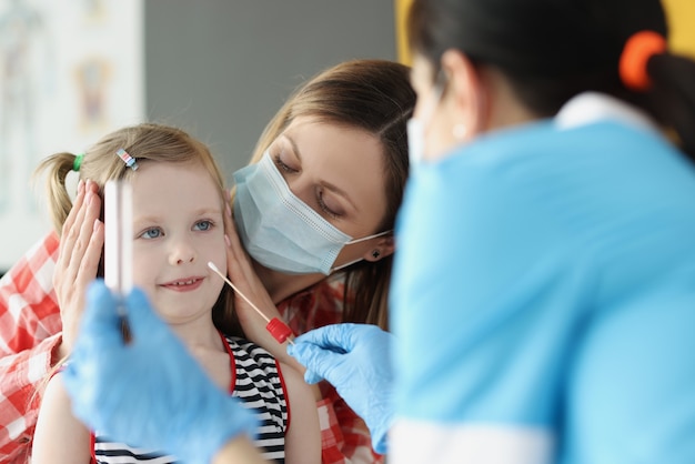 Доктор в защитной маске делает мазок ватной палочкой маленькой девочке в клинике