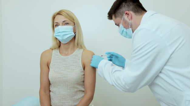 病院の女性患者にワクチン注射を行う保護マスクの医師。科学機器を備えた診療所でのコロナウイルス療法での生化学の医療ショット。