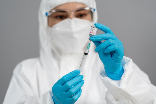 コロナウイルス（Covid-19）ワクチンボトルを注射器注射薬に引き込む保護PPEスーツの医師