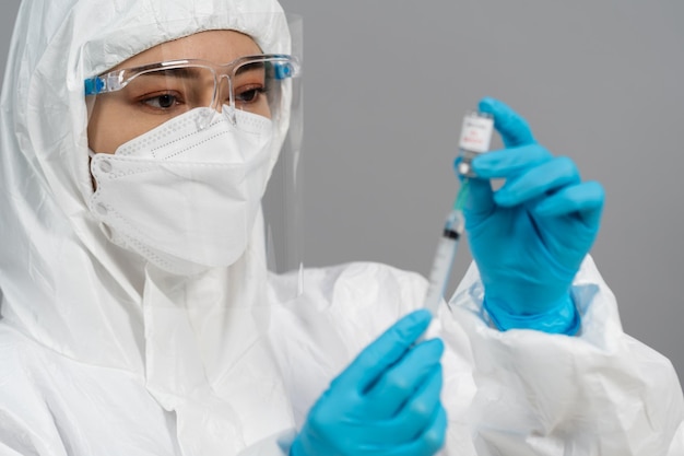 doctor in protective ppe suit drawing coronavirus (covid-19) vaccine bottle into syringe injection medicine