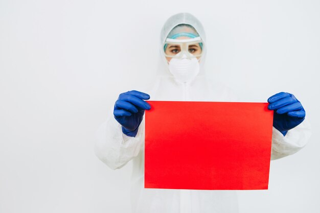 Doctor in protective suit and gloves holds sheet of red paper on white. doctor is battling coronavirus. COVID 19 virus.