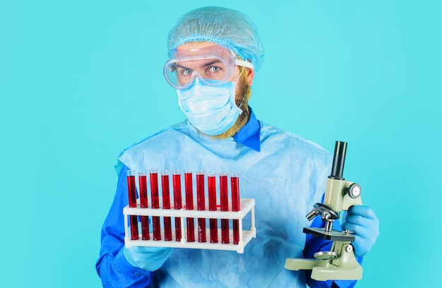 Doctor in protective suit, mask and glasses with test tubes with blood test and microscope.