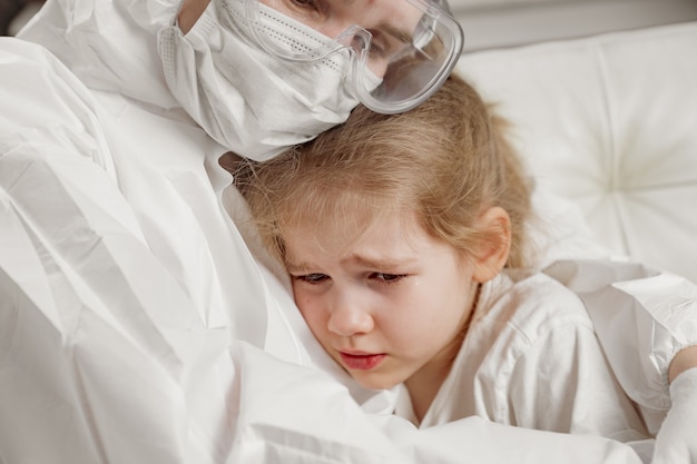 The doctor in a protective suit, mask, gloves and glasses calms down, hugs a little sick girl.