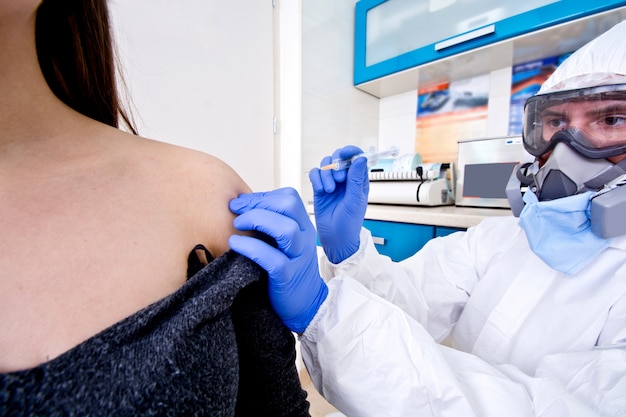 Doctor in protective suit uniform and mask giving a vaccine to female patient.