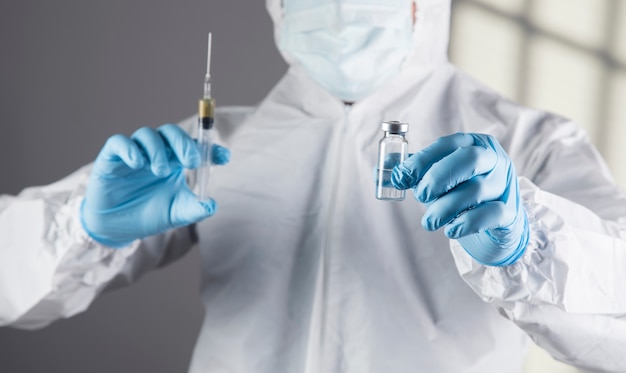 Doctor in protective uniform holds a syringe and ampoule on a gray scene