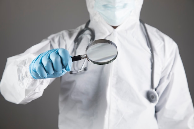 Doctor in protective uniform looks at a pill with a magnifying glass on a gray scene