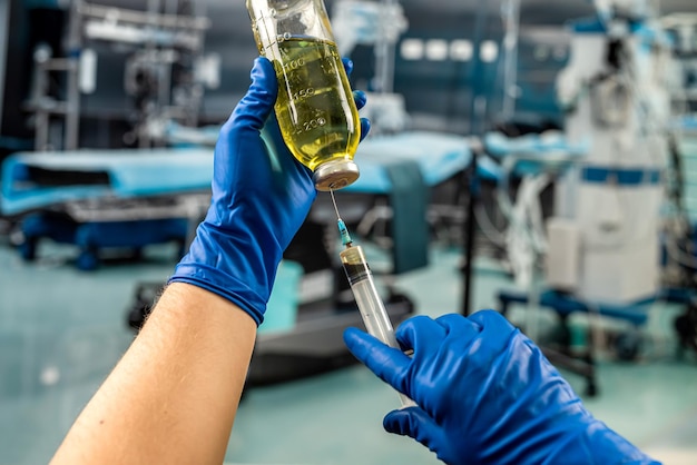 青い手袋をはめた医師の手は、病院の手術室で手術の準備をしている薬のワクチンまたは麻酔バイアルのボトルと注射器を持っています