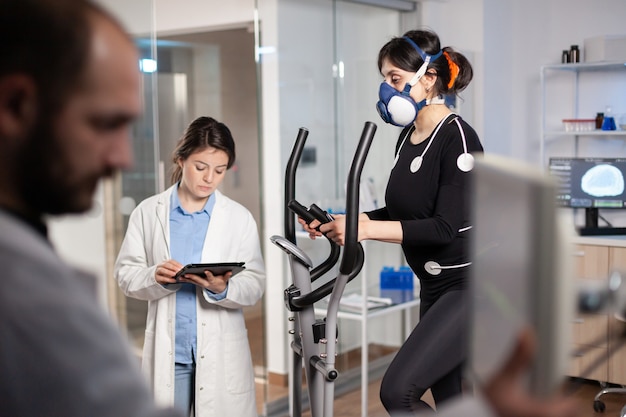 Scienziato medico che tiene in mano un tablet pc e controlla i dati ecg visualizzati sui monitor di laboratorio. ricercatore scientifico di laboratorio che misura la resistenza dell'atleta, la frequenza cardiaca utilizzando elettrodi e maschera.