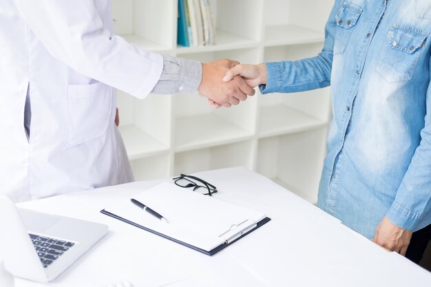 Il dottore stringe la mano all'ufficio medico con paziente, indossa occhiali, stetoscopio e cappotto di laboratorio.