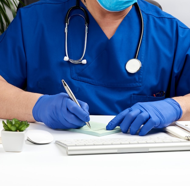 Foto medico seduto a un tavolo bianco in uniforme blu e guanti in lattice, specialista scrive una prescrizione per una farmacia