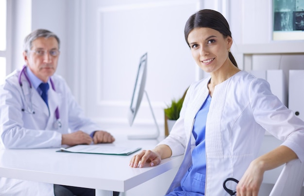 A doctor smiling at the camera with her male colleage in the back of the consulting room in hospital