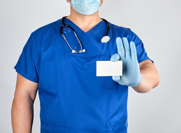 Medico in guanti di lattice sterili e uniforme blu tiene un biglietto da visita bianco vuoto