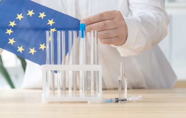 Doctor takes Flag of European Union near test tube, ampoule and syringe, concept of global vaccination.