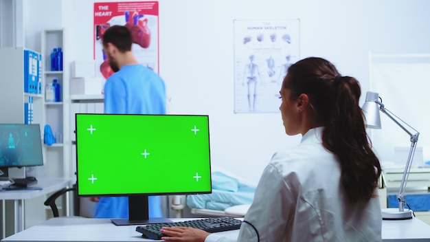 의사는 크로마 키 녹색 화면으로 컴퓨터에 메모를 하고 병원 캐비닛에는 파란색 유니폼을 입은 간호사가 있습니다. 환자 d를 확인하기 위해 클리닉 캐비닛에 빈 공간이 있는 모니터에서 작업하는 흰색 코트의 메딕