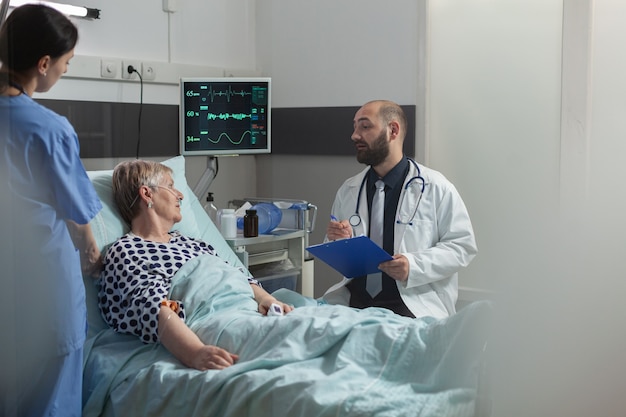 Doctor talking with elderly patient sitting next to bed in hospital room, giving expertise for treatment