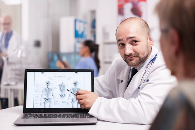 Doctor telling senior patient she needs back surgery during radiography examination in hospital office. Nurse in blue uniform holding x-ray in the background. Elderly physician in clinic corridor.