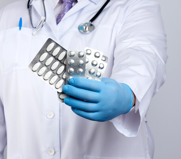 Doctor therapist is dressed in a white uniform coat and blue sterile gloves is standing and holding a stack of pills in blister packs