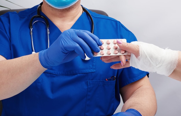 Doctor in uniform and blue latex medical gloves sits at a table and examines a patient with a hand injury