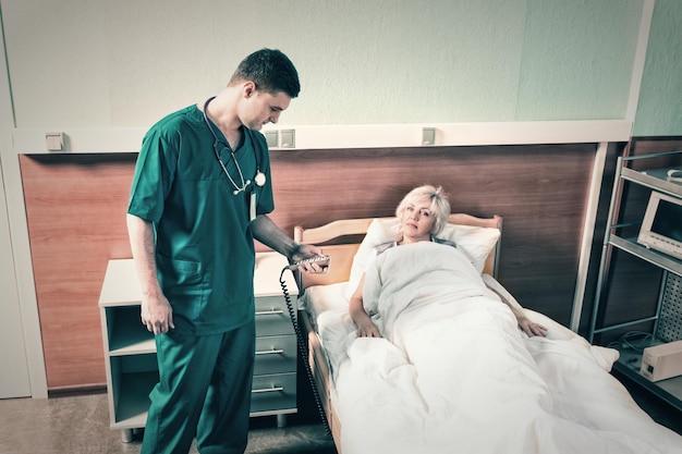 Doctor in uniform with phonendoscope on his neck regulates a hospital bed of his female patient using a special console in the hospital ward. Healthcare concept