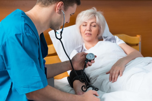 Doctor in uniform with phonendoscope is measuring the arterial pressure of his female patient using a medical equipment, who is lying in the bed in the hospital ward. Healthcare concept