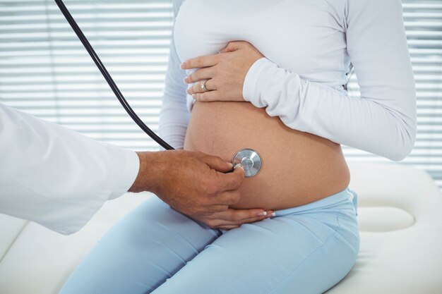 Doctor using stethoscope while examining pregnant woman in clinic