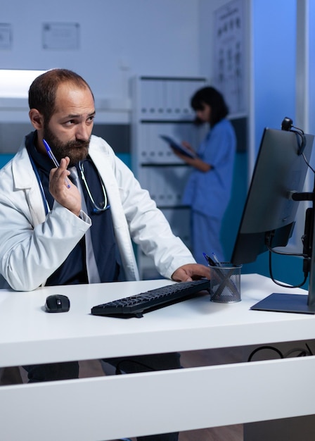 医師がコンピューターでビデオ通話を使用してリモート通信を行い、オフィスで遅くまで働いています。夜間の緊急遠隔医療相談のためのオンラインビデオ会議で患者と話している医療従事者。