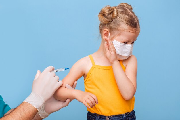 A doctor vaccinates a little blonde girl in a medical mask