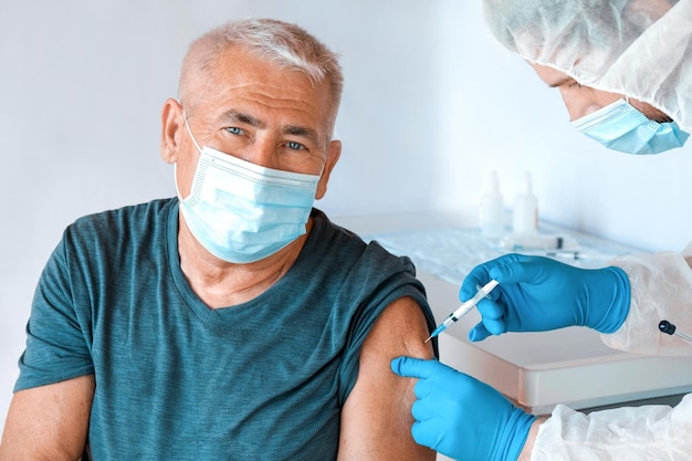 Доктор вакцинирует пожилого человека в клинике. Вакцинация пожилых людей. Доктор делает инъекцию вакцины против коронавируса COVID 19 взрослому мужчине в маске. Защита старшего пациента. Старик в больнице