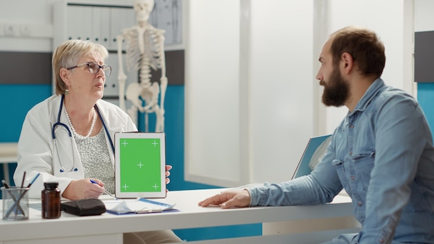 キャビネット内のデジタルタブレットでグリーンスクリーンを垂直に保持している医師。健康診断の人々は、孤立したクロマキーの背景と空白のコピースペースが表示されたモックアップテンプレートを見て訪問します。