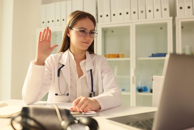 Doctor waving at laptop screen in clinic room remote medical consultation online concept