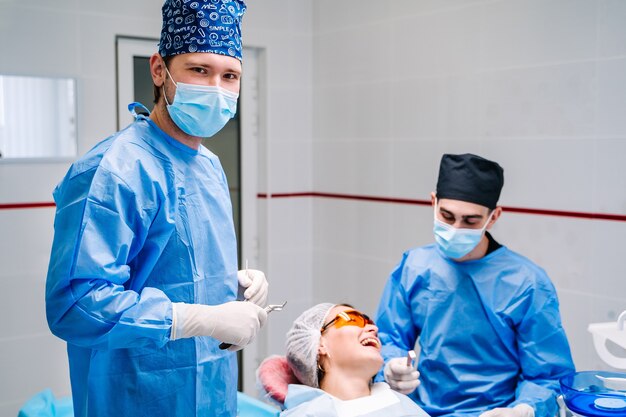 Medico che indossa camice blu e maschera che ti invita all'ufficio del dentista e che guarda l'obbiettivo con sedia stomatologica e paziente nel