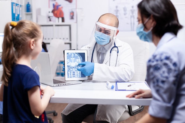 Doctor wearing face mask pointing at x-ray on tablet pc during child consultation