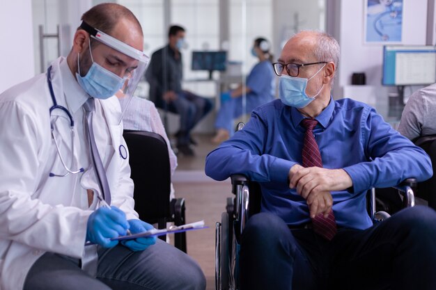 Doctor wearing face mask and visor against infection with coronavirus talking with disabled senior man in waiting area sitting on wheelchair