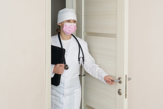 聴診器で医療用フェイスマスクを身に着けている医者は部屋に入るドアを開ける