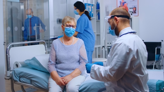 Doctor wearing a visor checking old retired senior woman lungs radiography in modern private hospital clinic. Healthcare facility during COVID-19 global pandemic, wearing protective equipment. Medical